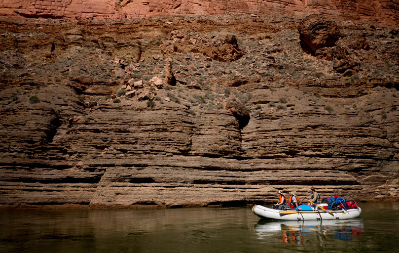 A raft floats down the river in front of a wall containing millions of years of sedimentary rock