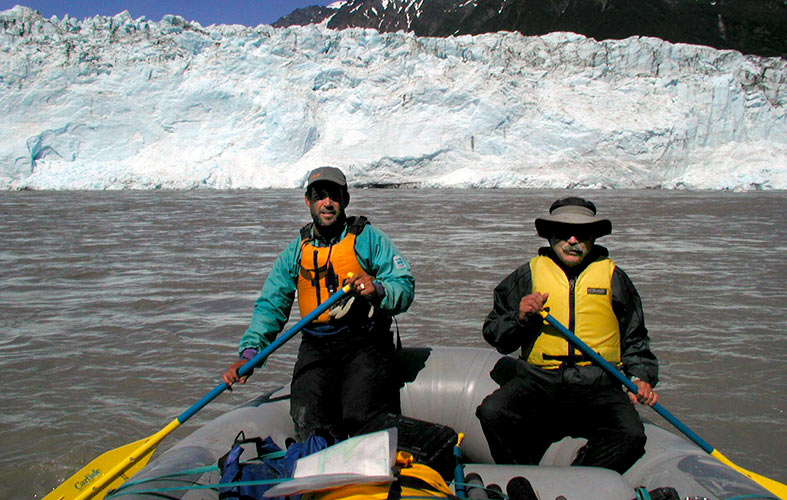 two men paddle a boat with a glacier visible in the background