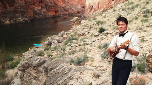 A man dressed in a dress shirt and bowtie plays the a ukelele on the shores of the Colorado River